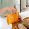 Person holding an orange cloth with white patterns over a wooden cutting board with bread slices.