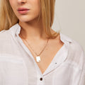 Person wearing a gold necklace with a tag pendant on a plain background