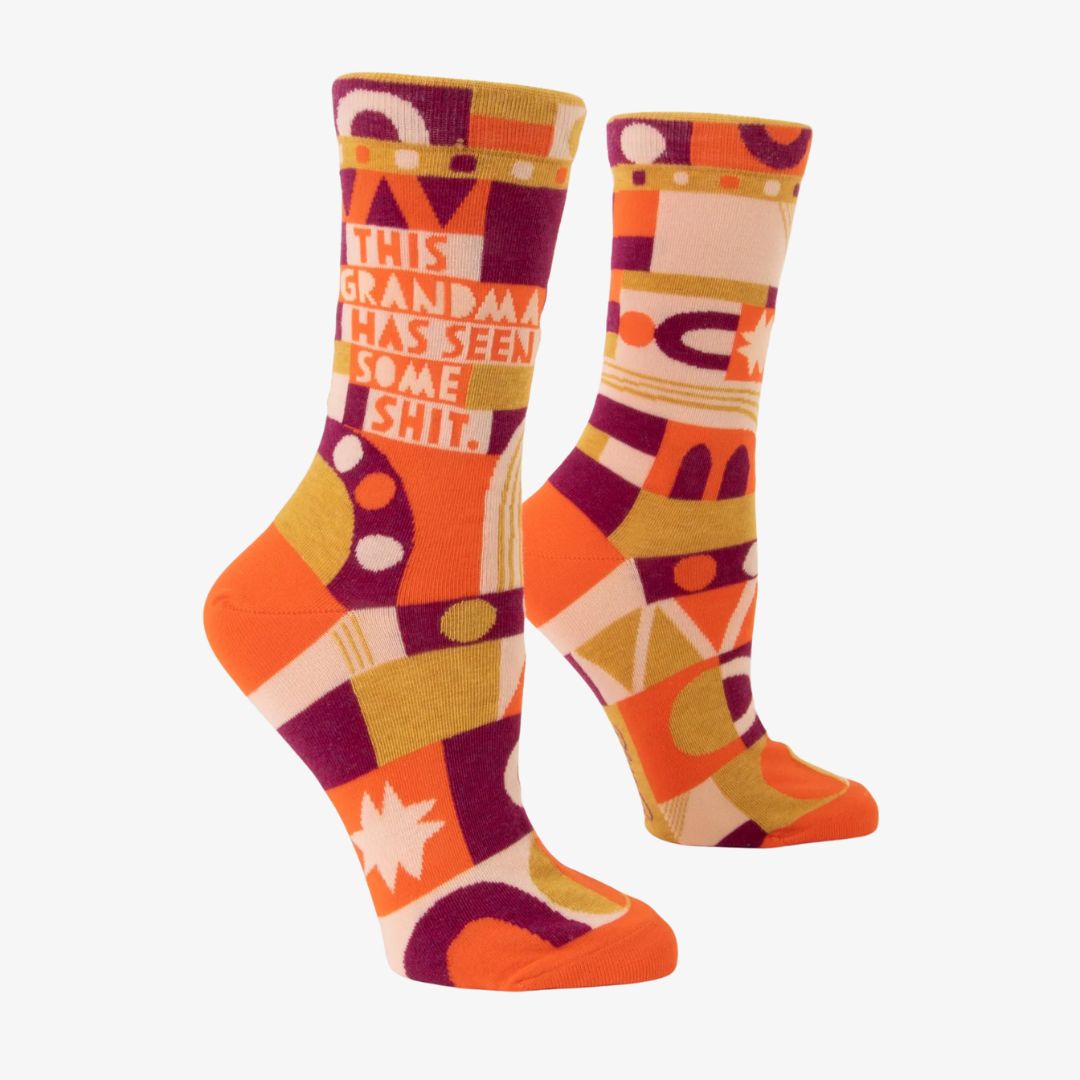 A pair of women's crew socks with a bold, abstract pattern in shades of orange, yellow, pink, and purple. Text on one sock reads "THIS GRANDMA HAS SEEN SOME SHIT."