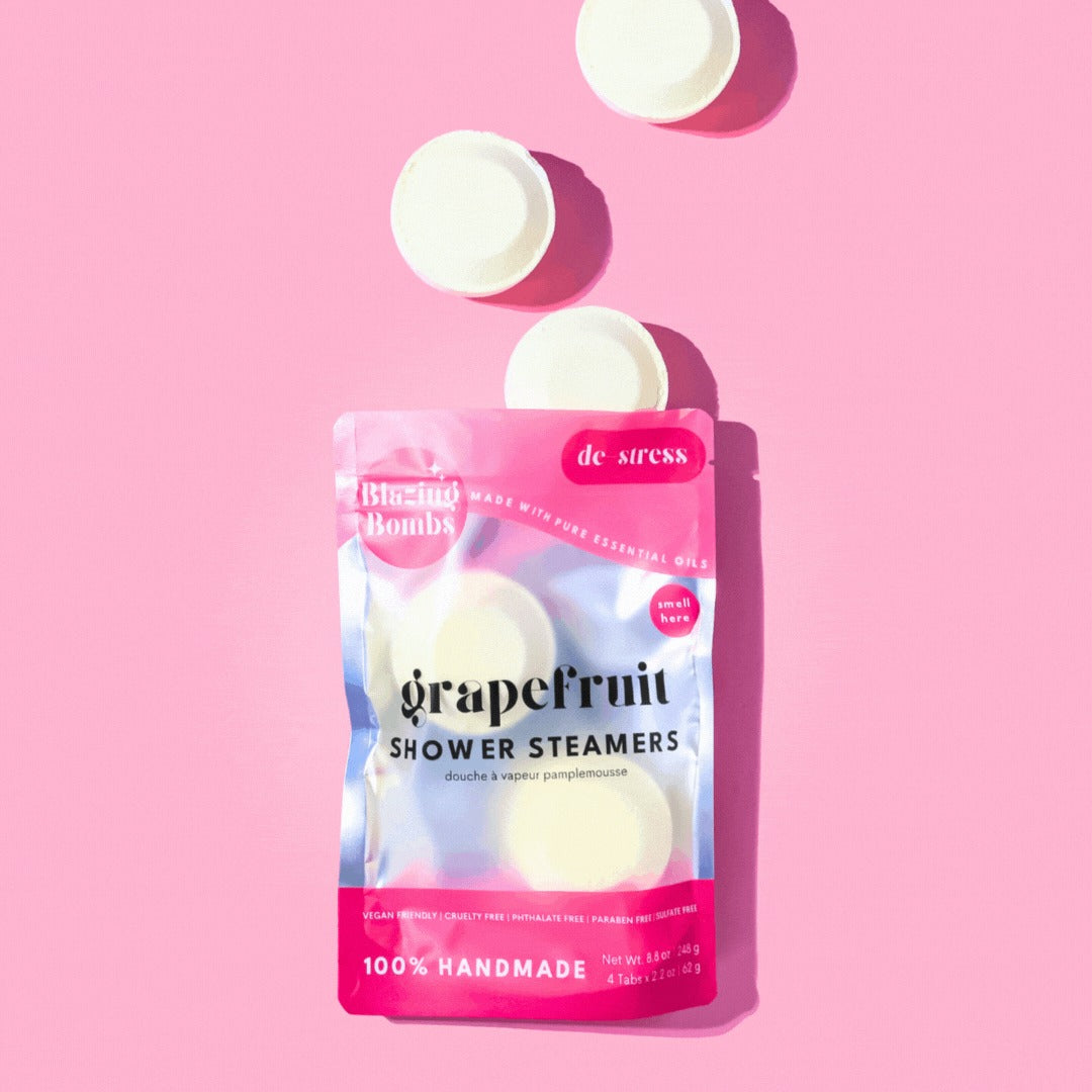 A vibrant, high-angle flat lay shot on a pink background features a shiny, pink and silver pouch of Blazing Bombs Grapefruit Shower Steamers. The pouch is labeled "de-stress" and contains four visible white, round steamer pucks. Three additional white pucks are floating above the bag, casting shadows on the pink surface. The label highlights that the product is 100% Handmade, Vegan Friendly, and made with Pure Essential Oils.