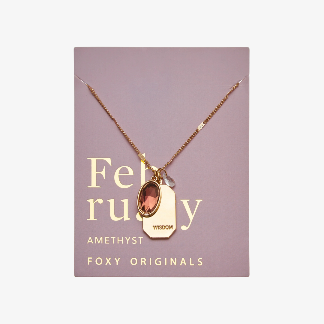 A gold necklace with an amethyst stone pendant. The pendant also has the word "WISDOM" engraved on it. The necklace is displayed on a purple card with the month "February" and the brand name "Foxy Originals."
