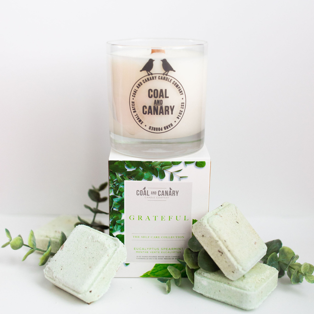 Grateful Candle, a glass jar candle with a wood wick, surrounded by eucalyptus and bath soaks.