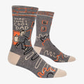 A pair of grey men's crew socks with orange, white, and black abstract lines. One sock features a cartoon drawing of a man with an orange background and the text "HERE COMES COOL DAD" and "HIGH FIVE".