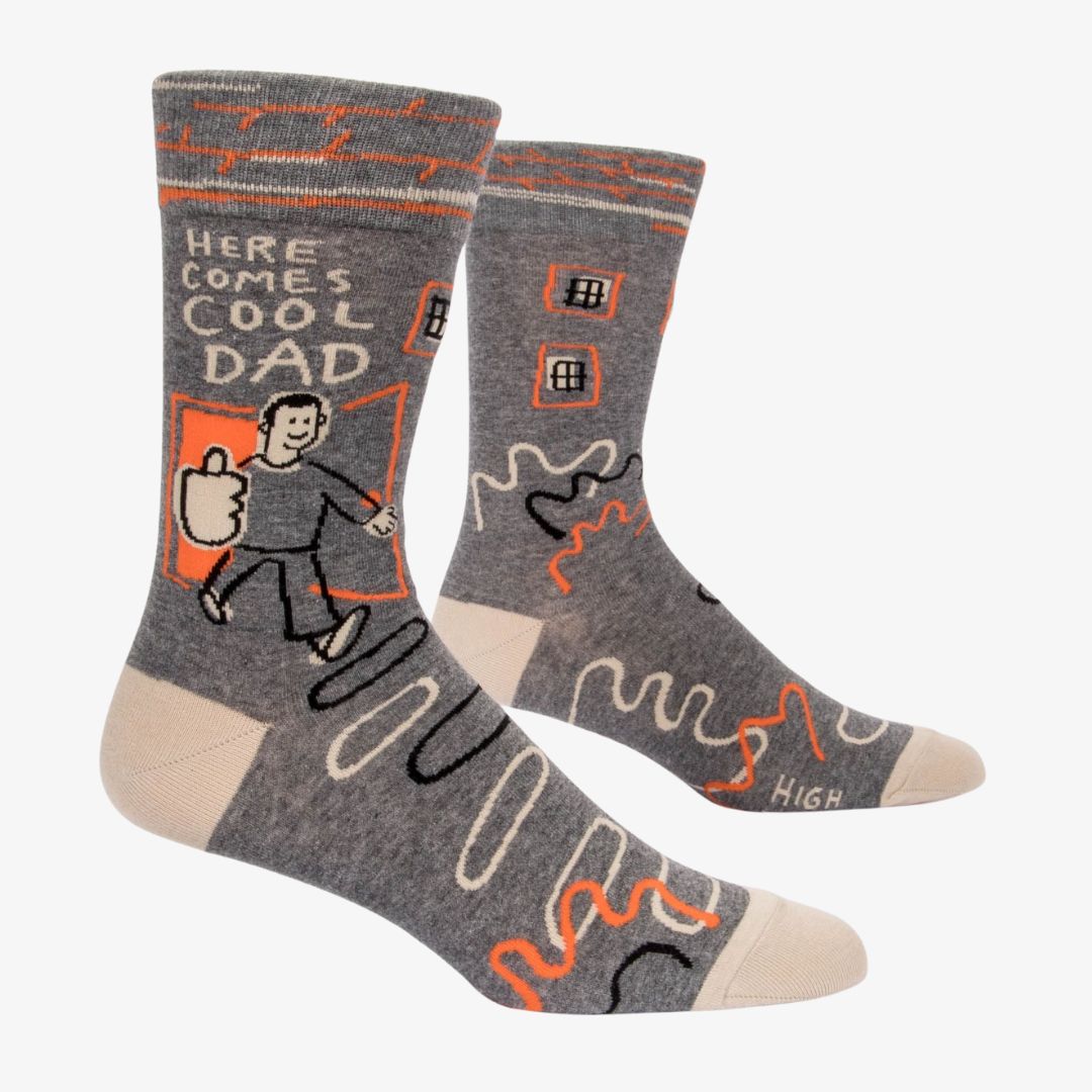 A pair of grey men's crew socks with orange, white, and black abstract lines. One sock features a cartoon drawing of a man with an orange background and the text "HERE COMES COOL DAD" and "HIGH FIVE".