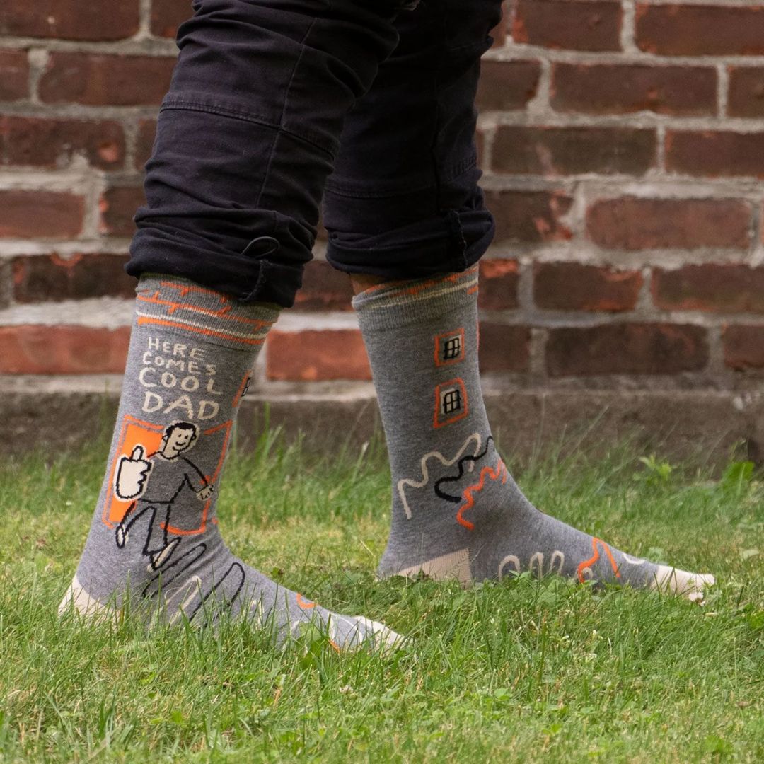A pair of grey men's crew socks with orange, white, and black abstract lines. One sock features a cartoon drawing of a man with an orange background and the text "HERE COMES COOL DAD" and "HIGH FIVE".