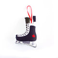 A close-up, high-angle flat lay shot on a plain white background features a soft, plush Christmas ornament shaped like a hockey skate. The skate is black and white, with an illustration of a red maple leaf on the side of the boot. The blade of the skate is grey and white. A red ribbon loop is attached to the top for hanging.