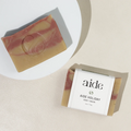 A bar of Aide Bodycare Holiday Soap, featuring a layered light yellow and red color and a paper label with the 'aide' logo and 'Aide Holiday' text, displayed on a light-colored surface with a shadow overlay. The soap is natural and handmade, available in Ottawa, Ontario, Canada.