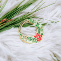 A gold-colored pin shaped like a Christmas wreath. The wreath is decorated with green leaves, red berries, and a red poinsettia flower.