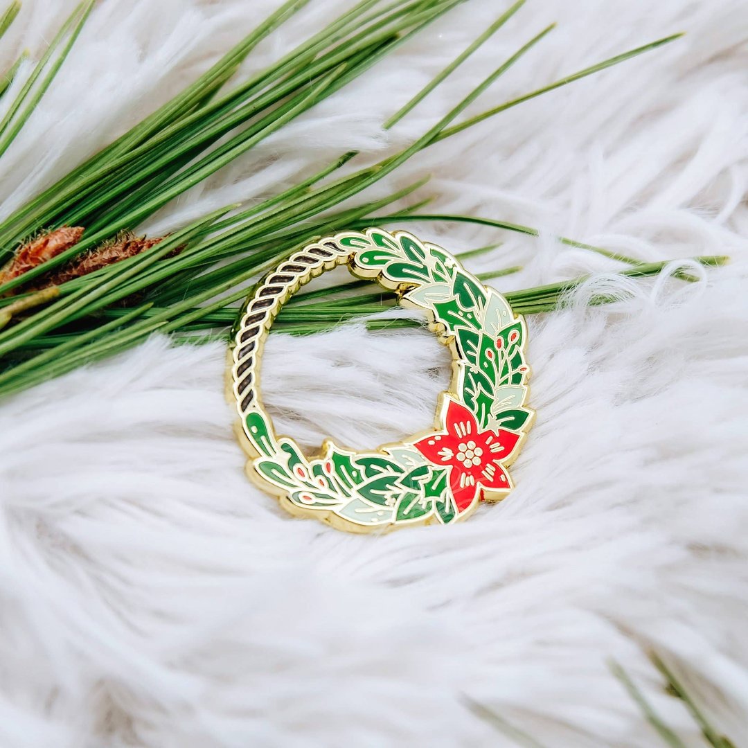 A gold-colored pin shaped like a Christmas wreath. The wreath is decorated with green leaves, red berries, and a red poinsettia flower.