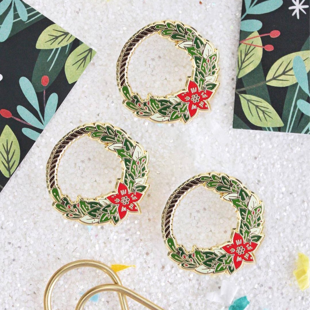 A gold-colored pin shaped like a Christmas wreath. The wreath is decorated with green leaves, red berries, and a red poinsettia flower.