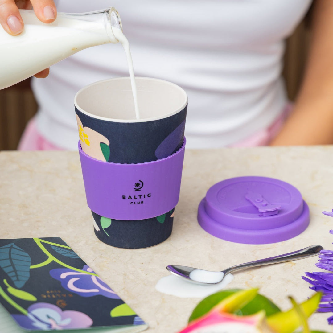 Person pouring milk into a coffee cup with a purple sleeve, surrounded by colorful items on a table.