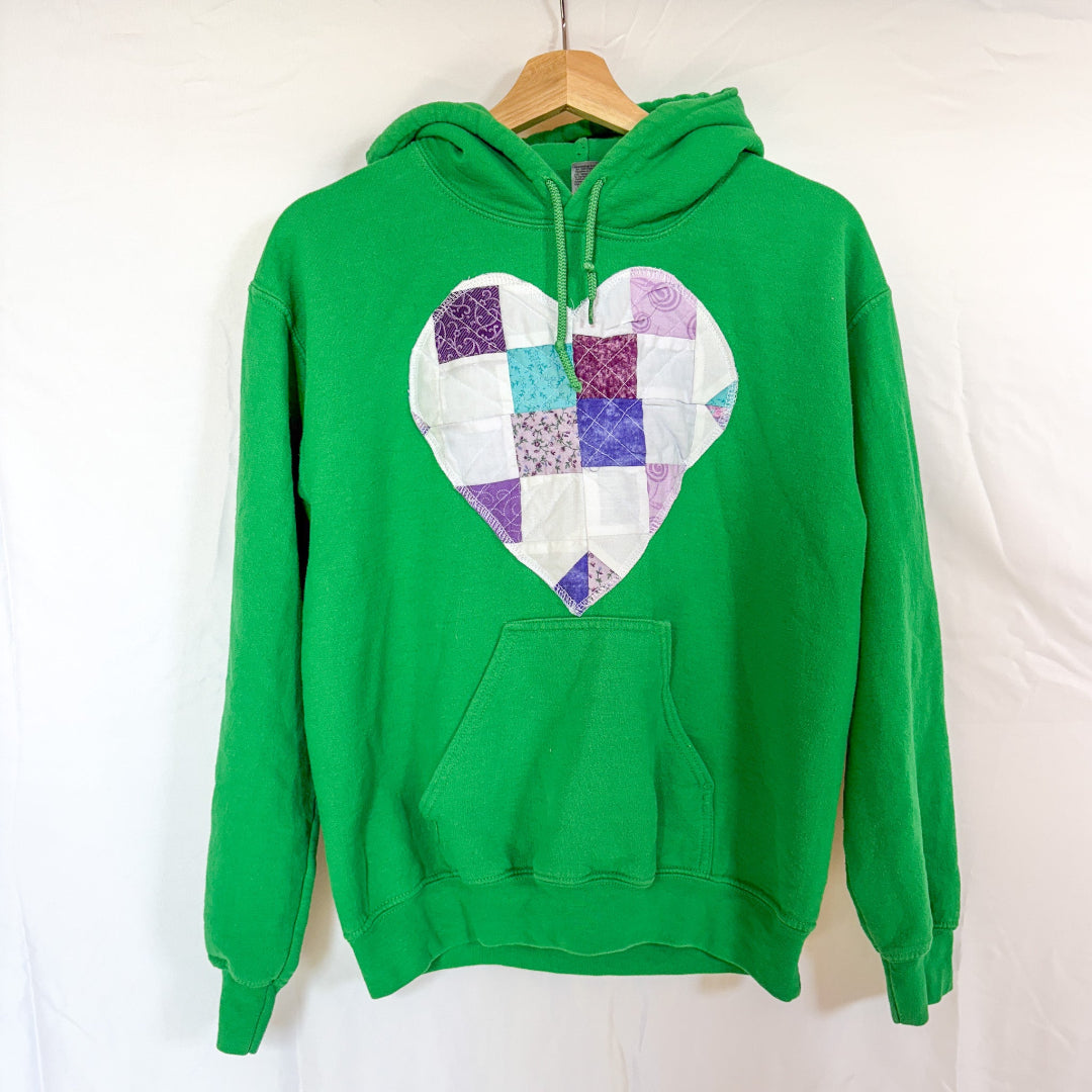 A bright green Gildan hoodie, size X-Small, hanging on a wooden hanger against a white background. The front of the hoodie is visible, with a large, heart-shaped quilted patchwork sewn onto the chest. The heart is made of various fabric scraps in shades of blue, purple, and white with different patterns.