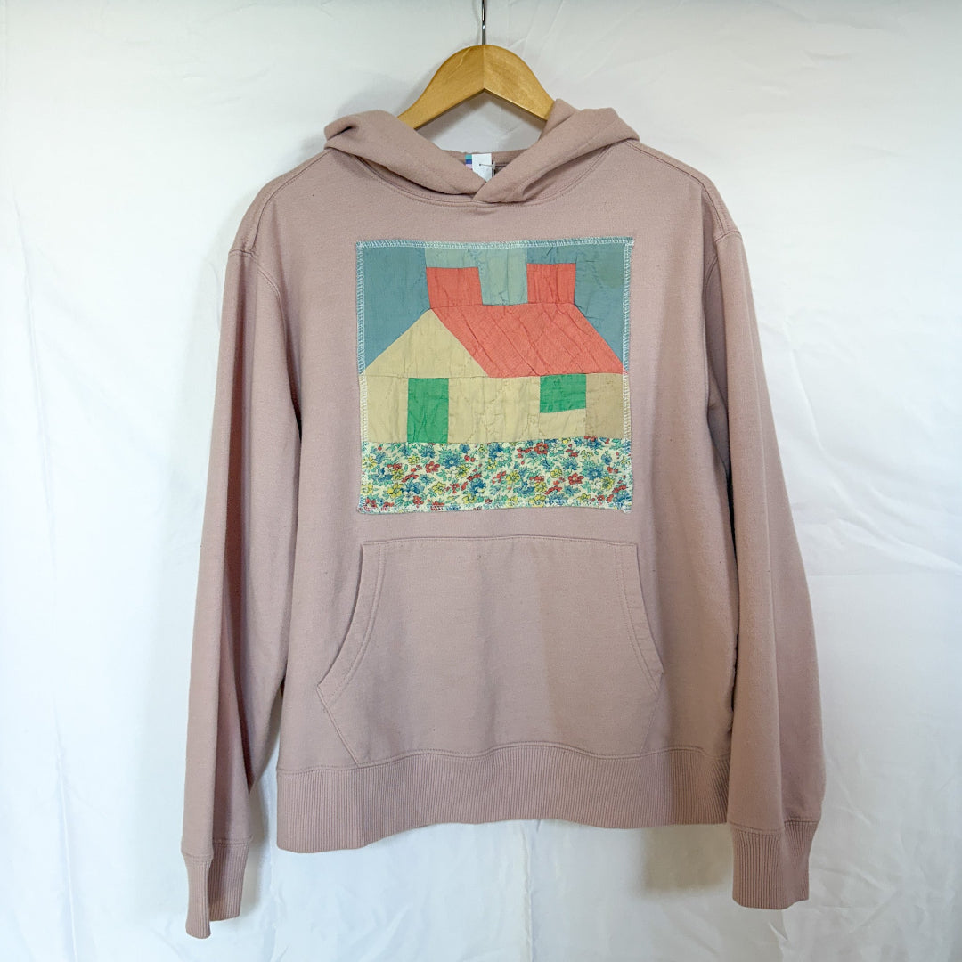 A light pink Janet Jackson hoodie, size Medium, hanging on a wooden hanger against a white background. The front of the hoodie is visible, with a large, square quilted panel sewn onto the chest. The patch depicts a house with a red roof, a light blue sky, green doors, and a base of multicoloured floral fabric.