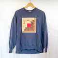 A navy blue crewneck sweatshirt, size X-Small, hanging on a wooden hanger against a white background. The front of the sweatshirt is visible, with a square, quilted patchwork panel sewn onto the chest. The patch is made of various fabric scraps in warm tones of brown, beige, and red with floral and polka-dot patterns.