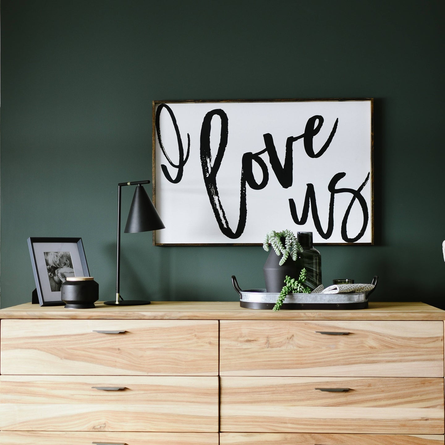 Wooden dresser with decorative items including a framed 'I love us' sign, lamp, and plants against a dark green wall.