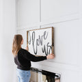 Person hanging a framed sign with 'I love us' text on a white wall.