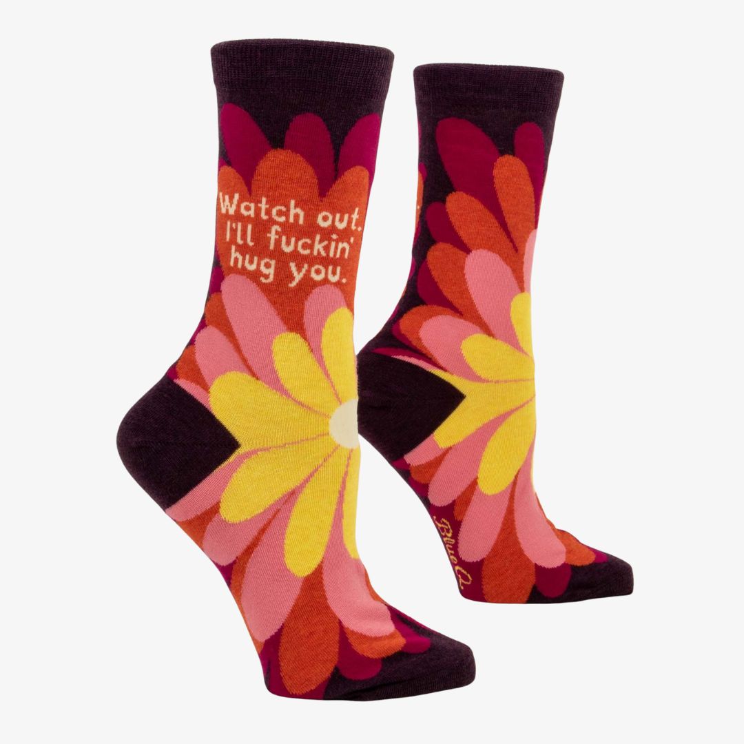A pair of women's crew socks with a dark purple background and a vibrant, large floral pattern in shades of red, fuchsia, pink, and yellow. Text on one sock reads "Watch out. I'll fuckin' hug you."