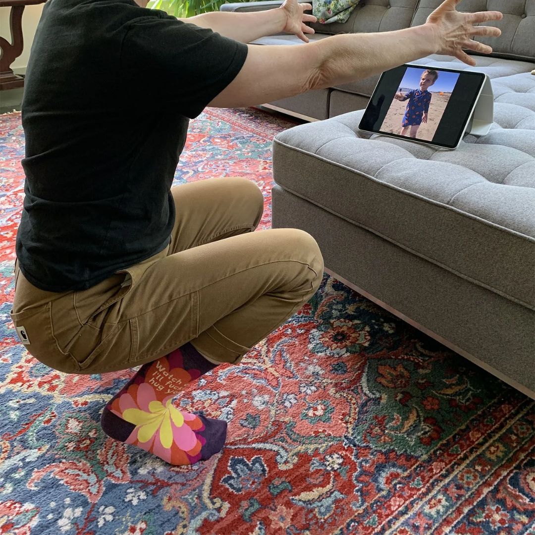 A pair of women's crew socks with a dark purple background and a vibrant, large floral pattern in shades of red, fuchsia, pink, and yellow. Text on one sock reads "Watch out. I'll fuckin' hug you."