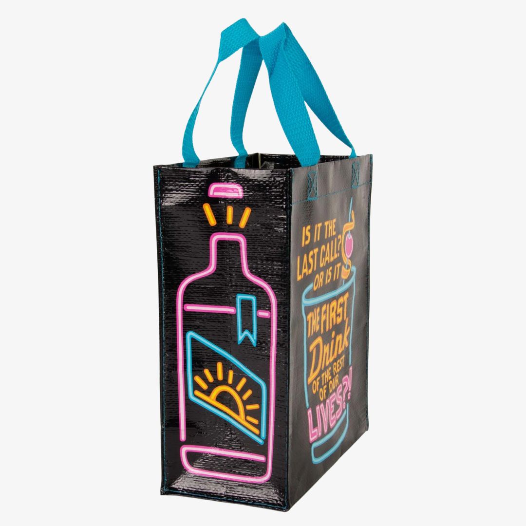 A black reusable tote bag with bright neon blue, pink, and yellow graphics. The front features a large cocktail glass with a cherry and straw, and text that reads "IS IT THE LAST CALL? OR IS IT THE FIRST DRINK OF THE REST OF OUR LIVES?!". On the left side, part of a neon bottle graphic is visible.