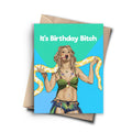A humorous birthday card featuring a cartoon drawing of Britney Spears from her performance at the 2001 MTV Video Music Awards, holding a yellow python around her neck. The text on the card reads, "It's Birthday Bitch." A brown kraft envelope is behind the card.