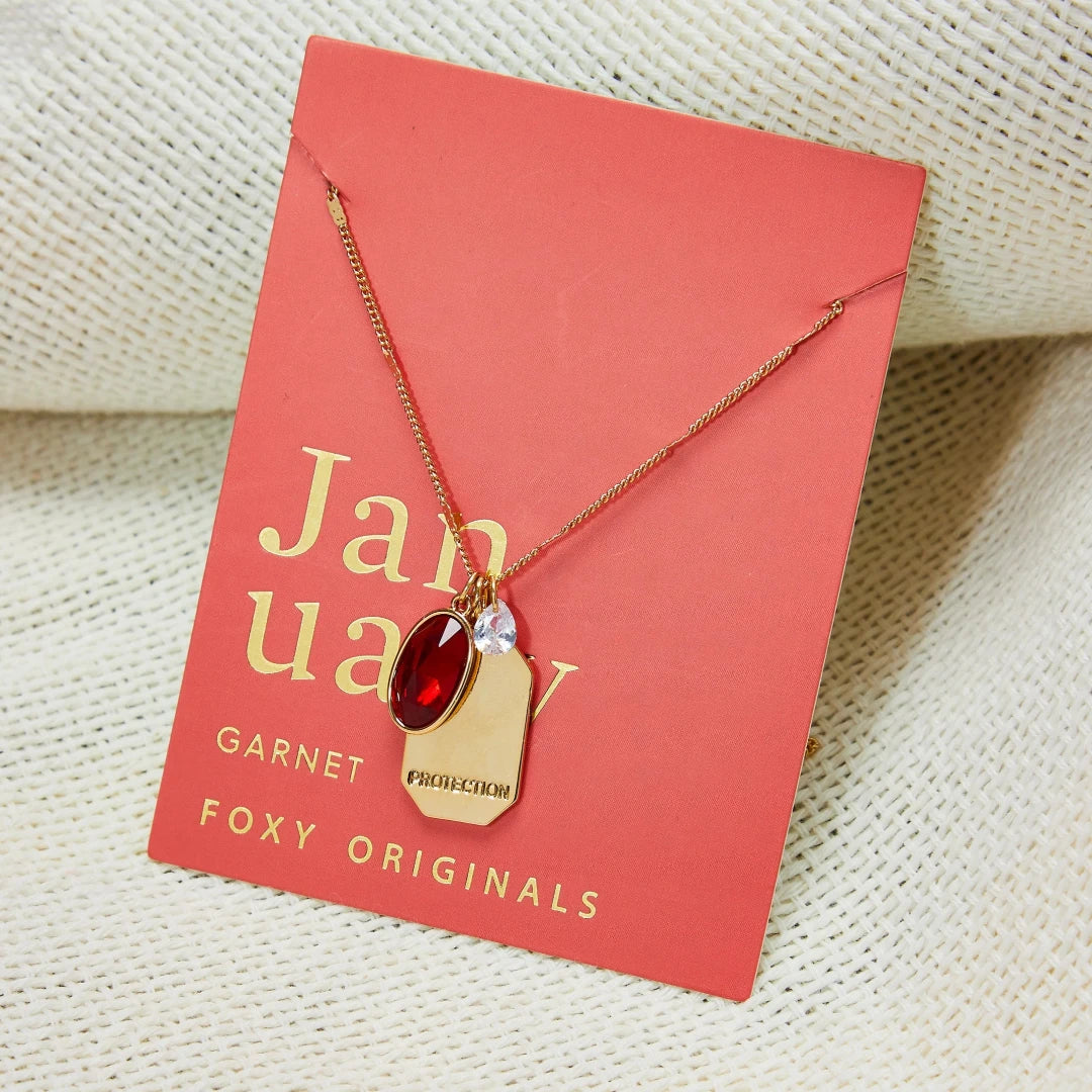 A gold necklace with a red garnet stone pendant. The pendant also has the word "Protection" engraved on it. The necklace is displayed on a red card with the month "January" and the brand name "Foxy Originals."