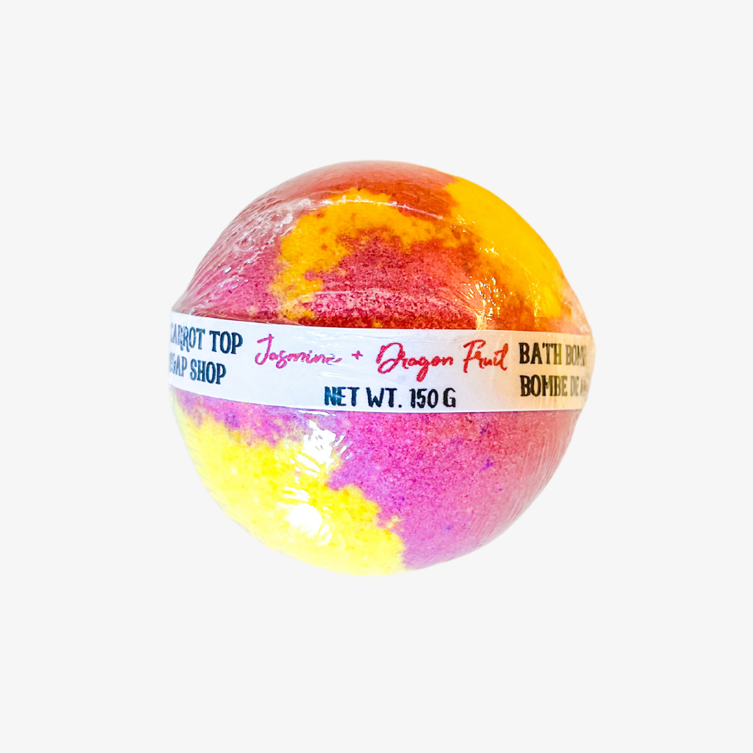 A round, multicolored bath bomb wrapped in plastic. The bath bomb has swirls of yellow, orange, and pink, and is labeled "Jasmine + Dragon Fruit" and "Net Wt. 150g".