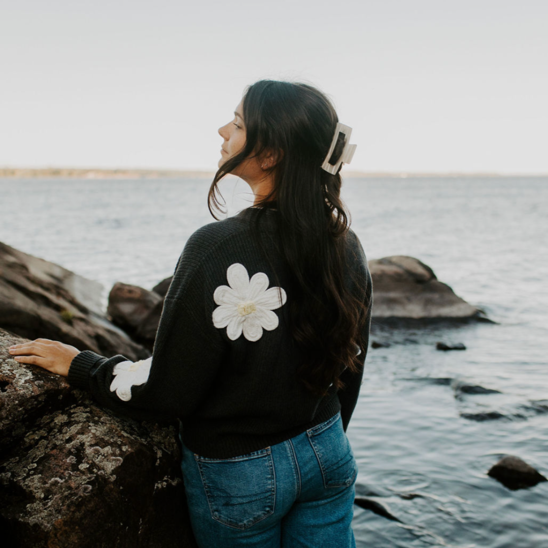 Person wearing a black jacket with a white flower design by a body of water.
