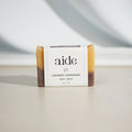 A bar of Aide Bodycare Lavender Lemongrass Soap, featuring a layered light yellow and dark brown color and a paper label with the 'aide' logo and 'Lavender Lemongrass' text, displayed on a light-colored surface with a shadow overlay. The soap is natural and handmade, available in Ottawa, Ontario, Canada.