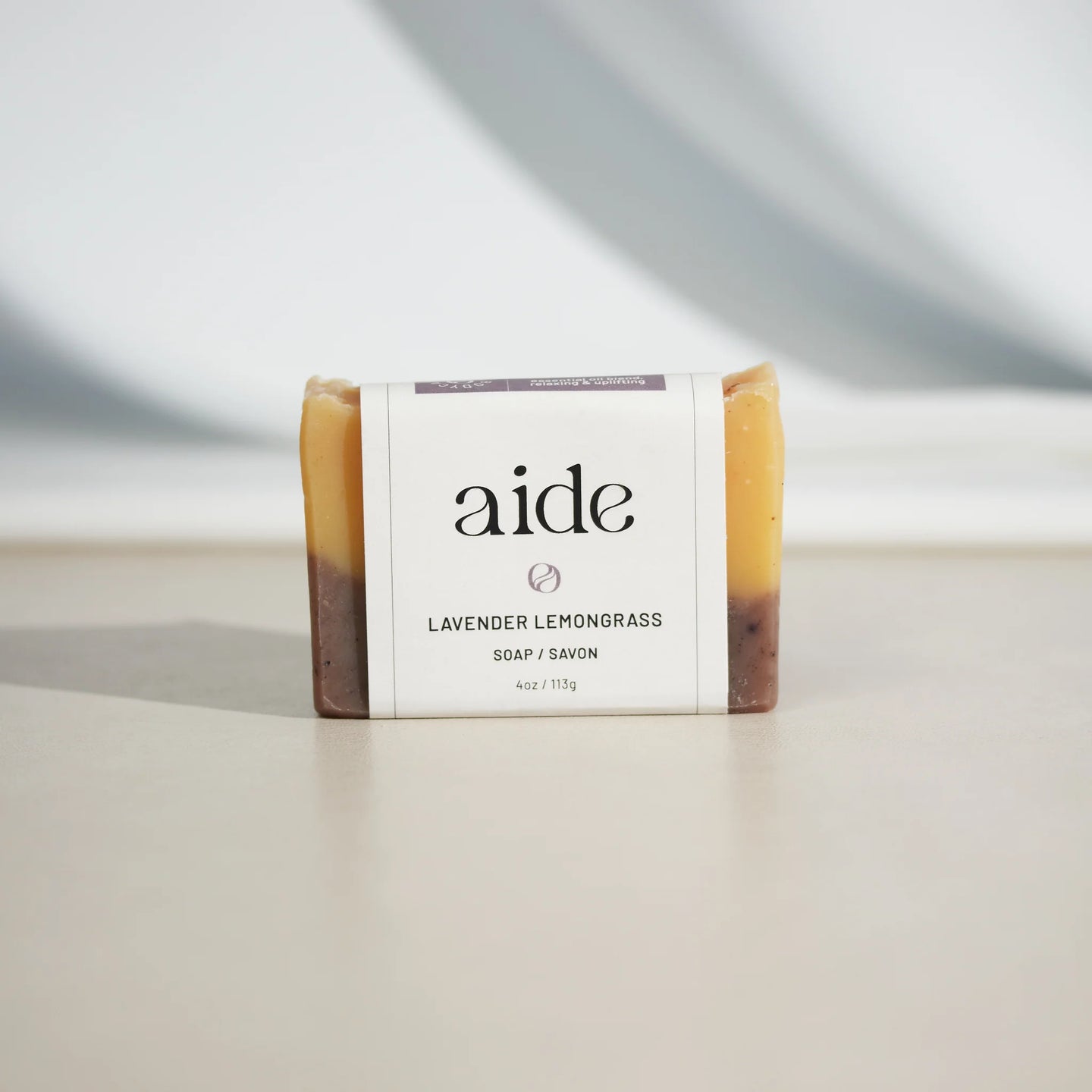A bar of Aide Bodycare Lavender Lemongrass Soap, featuring a layered light yellow and dark brown color and a paper label with the 'aide' logo and 'Lavender Lemongrass' text, displayed on a light-colored surface with a shadow overlay. The soap is natural and handmade, available in Ottawa, Ontario, Canada.