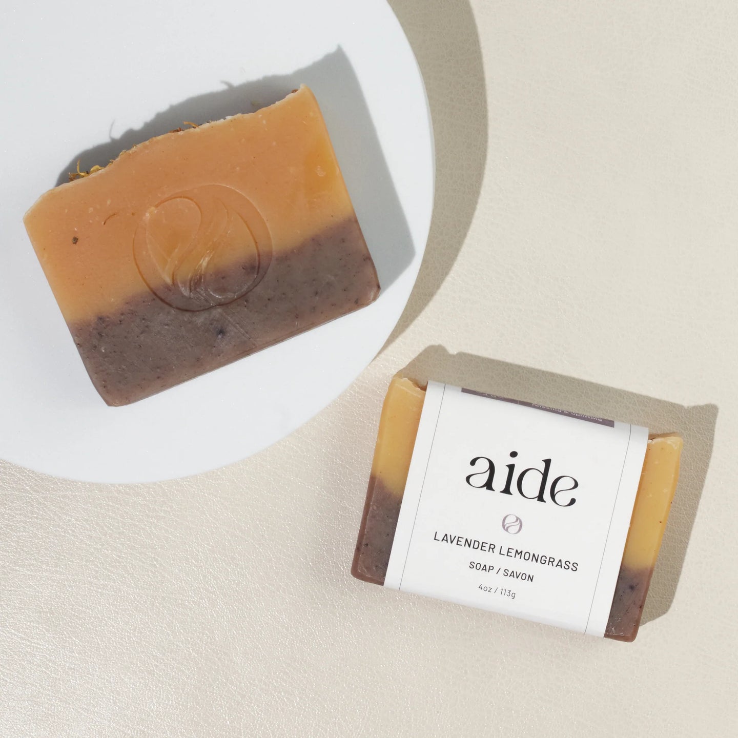 A bar of Aide Bodycare Lavender Lemongrass Soap, featuring a layered light yellow and dark brown color and a paper label with the 'aide' logo and 'Lavender Lemongrass' text, displayed on a light-colored surface with a shadow overlay. The soap is natural and handmade, available in Ottawa, Ontario, Canada.