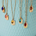 A collection of gold necklaces with colored gemstone pendants displayed on a light blue surface. Each pendant has a word engraved on it, such as "JOY," "FRIENDSHIP," "WISDOM," "HEALING," and "PROTECTION." The necklaces are arranged in a semicircle, with the pendants facing the viewer.