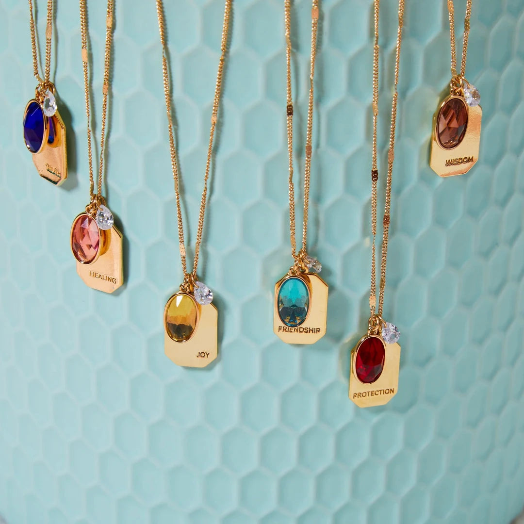 A collection of gold necklaces with colored gemstone pendants displayed on a light blue surface. Each pendant has a word engraved on it, such as "JOY," "FRIENDSHIP," "WISDOM," "HEALING," and "PROTECTION." The necklaces are arranged in a semicircle, with the pendants facing the viewer.