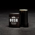 A black lip balm tube with a white label. The label reads "BUSH" and "BUSH BALM FOR LIPS & FACE." The balm is partially twisted up, revealing the product inside. The background is black.