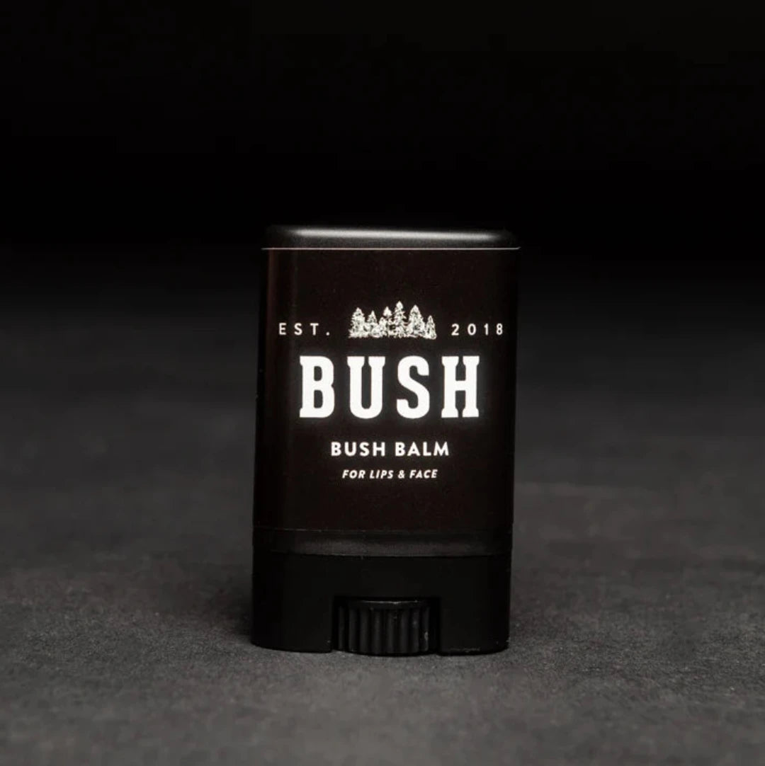 A black lip balm tube with a white label. The label reads "BUSH" and "BUSH BALM FOR LIPS & FACE." The balm is partially twisted up, revealing the product inside. The background is black.