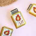 A hand holding a gold-colored enamel pin shaped like a matchbox. The matchbox has a lit match with a flame and the word "LIT" written on it.