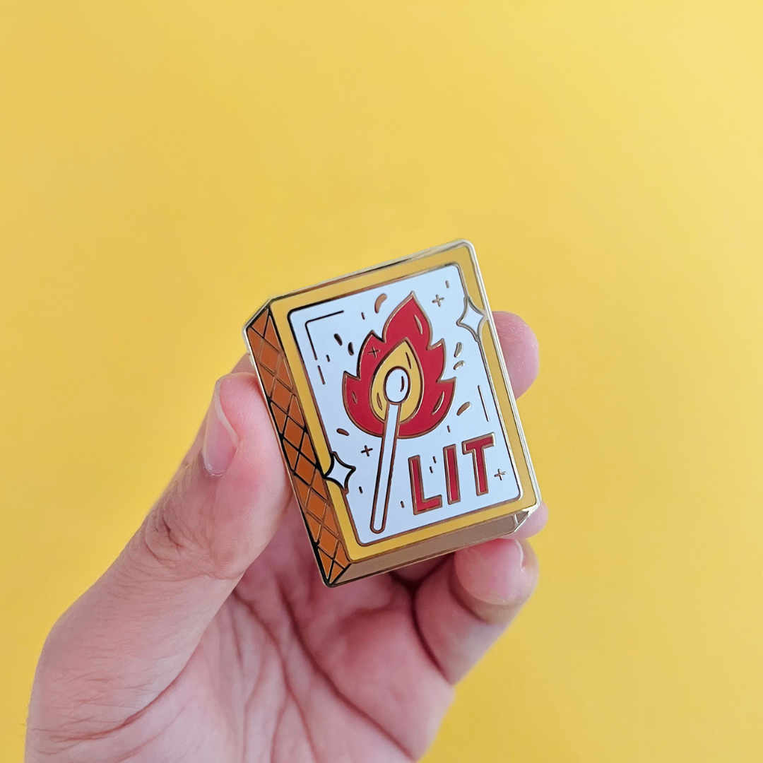 A hand holding a gold-colored enamel pin shaped like a matchbox. The matchbox has a lit match with a flame and the word "LIT" written on it.