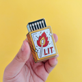 A hand holding a gold-colored enamel pin shaped like a matchbox. The matchbox has a lit match with a flame and the word "LIT" written on it.