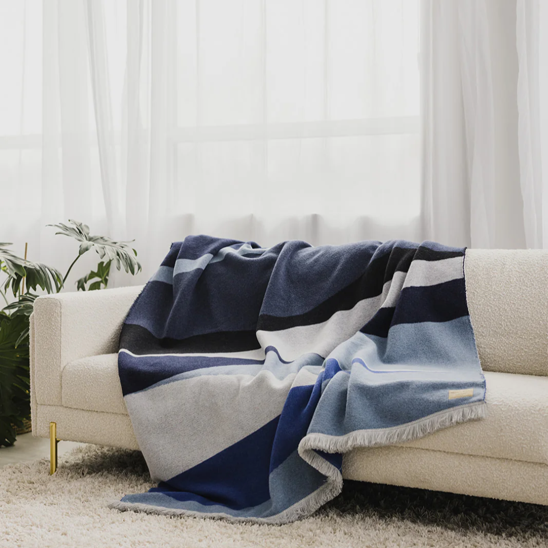 The River Reversible Blanket by Mini Tipi, featuring flowing waves of blue, grey, black, and white, held up by two hands. This reversible blanket is available in Ottawa, Ontario, Canada.