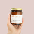Hand holding a jar of 'Maple Chai' candle against a pink background