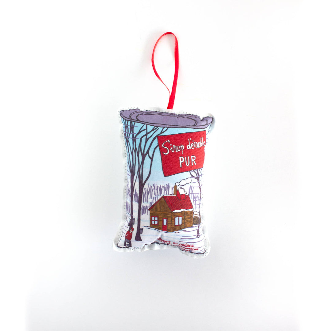 A close-up, high-angle flat lay shot on a plain white background features a soft, plush Christmas ornament shaped like a can of maple syrup. The can has a blue and red label with a cartoon winter scene showing a small wooden cabin among trees. A red oval banner in the center reads "Sirop d'érable PUR". The top of the can is colored purple and a red ribbon loop is attached for hanging.