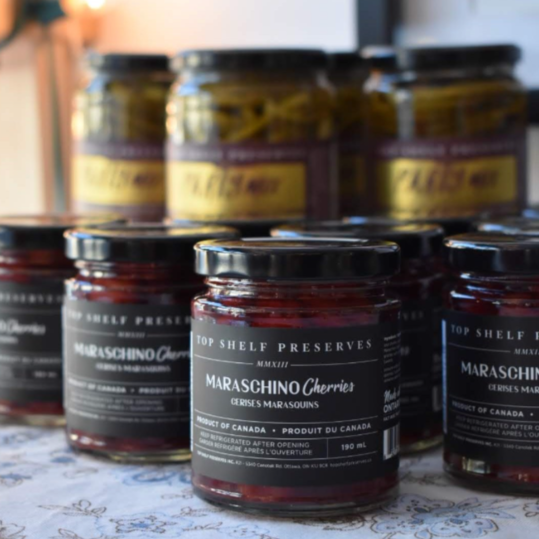 A jar of Maraschino Cherries from Top Shelf Preserves. The jar is filled with red cherries and has a black label with white text. The label includes the product name, "Product of Canada," and instructions to keep refrigerated after opening.