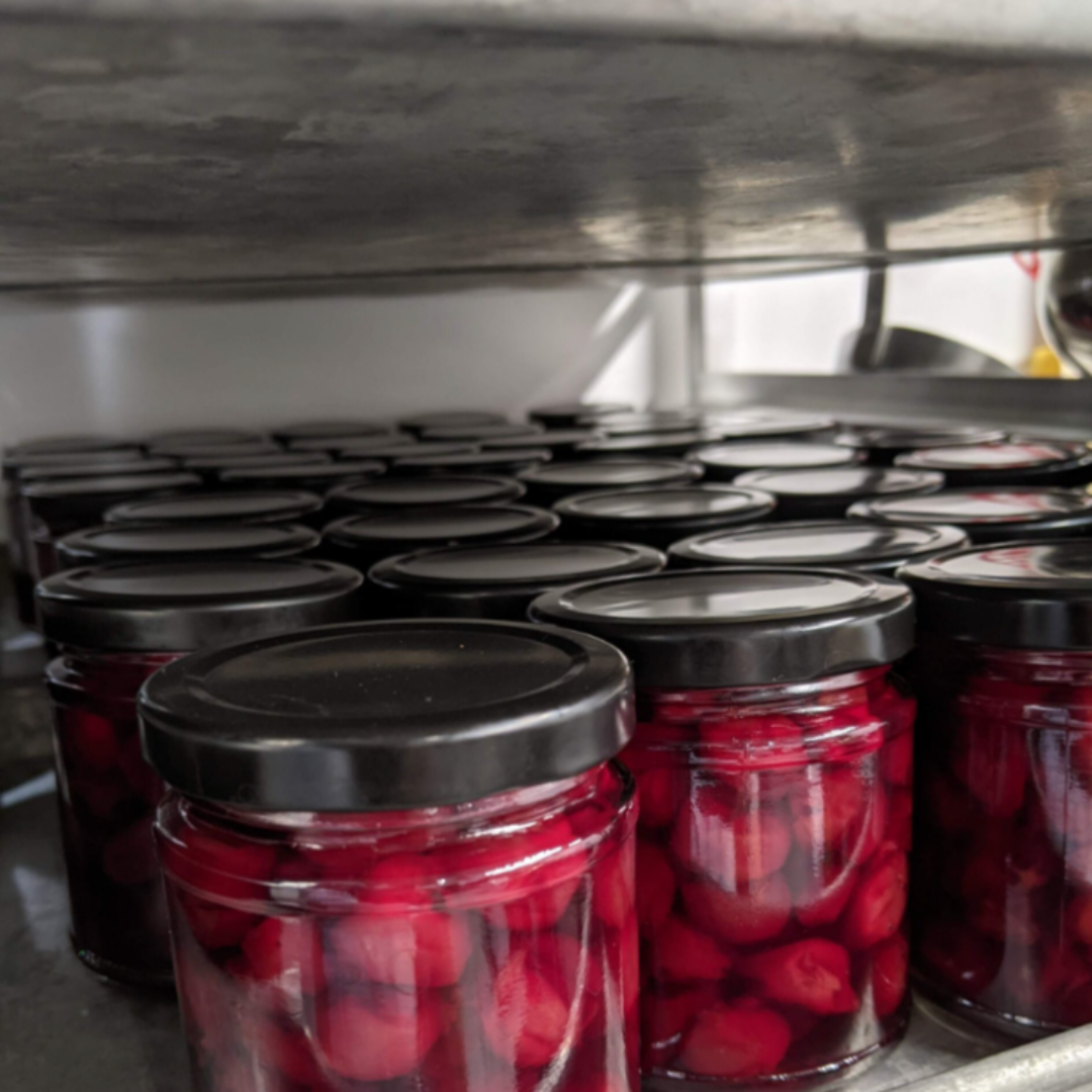 A jar of Maraschino Cherries from Top Shelf Preserves. The jar is filled with red cherries and has a black label with white text. The label includes the product name, "Product of Canada," and instructions to keep refrigerated after opening.