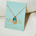 A gold necklace with an aquamarine stone pendant. The pendant also has the word "SERENITY" engraved on it. The necklace is displayed on a light blue card with the month "March" and the brand name "Foxy Originals."