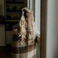 Person with long blonde hair wrapped in a plaid blanket, standing by a window.