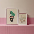 Two framed artworks on a pink tiled shelf against a pink wall.