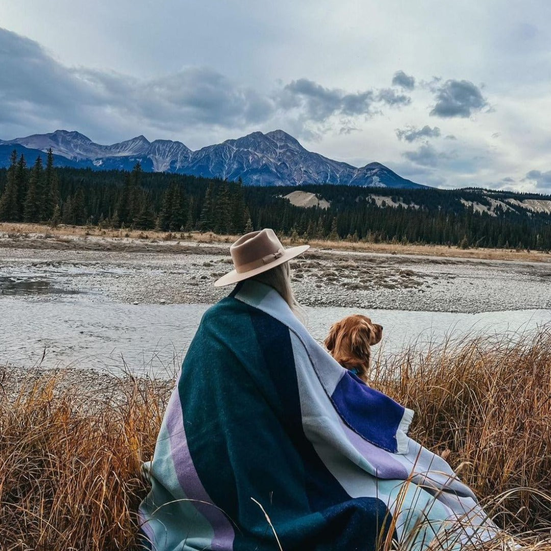 The Aurora Reversible Eco-Friendly Blanket by Mini Tipi, featuring flowing waves of green, purple, black, and white, draped on a woman and dog. This reversible and eco-conscious blanket is available in Ottawa, Ontario, Canada.