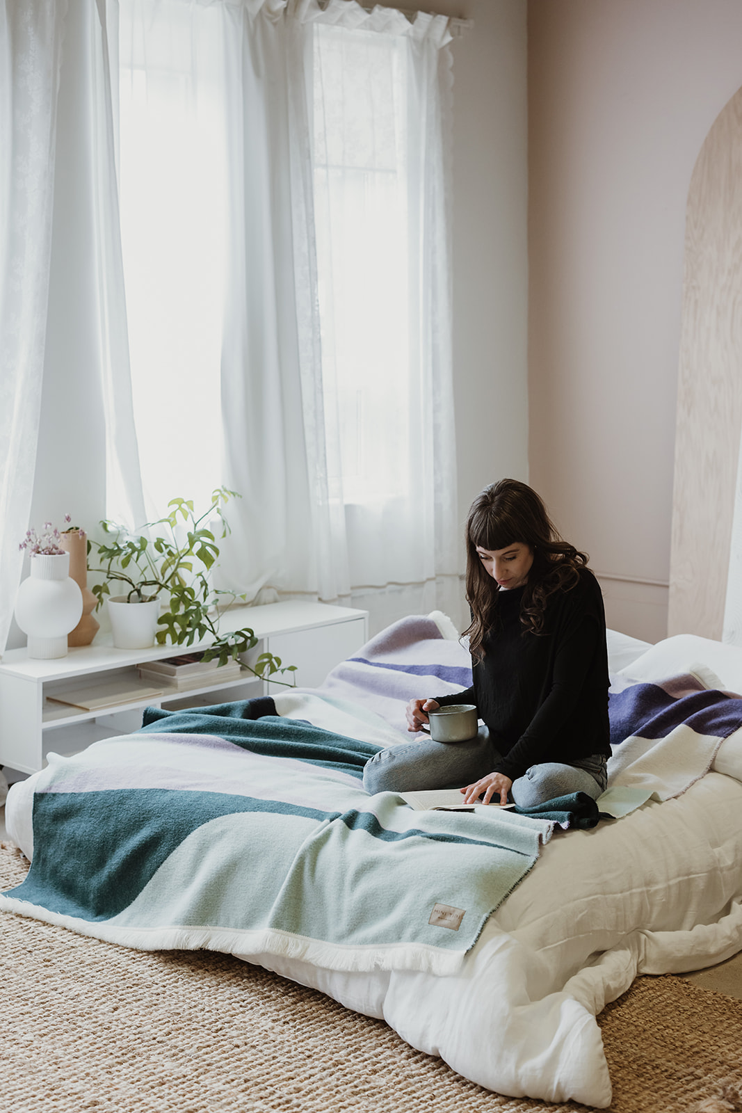 The Aurora Reversible Eco-Friendly Blanket by Mini Tipi, featuring flowing waves of green, purple, black, and white, laying on a bed with a person sitting on top. This reversible and eco-conscious blanket is available in Ottawa, Ontario, Canada.