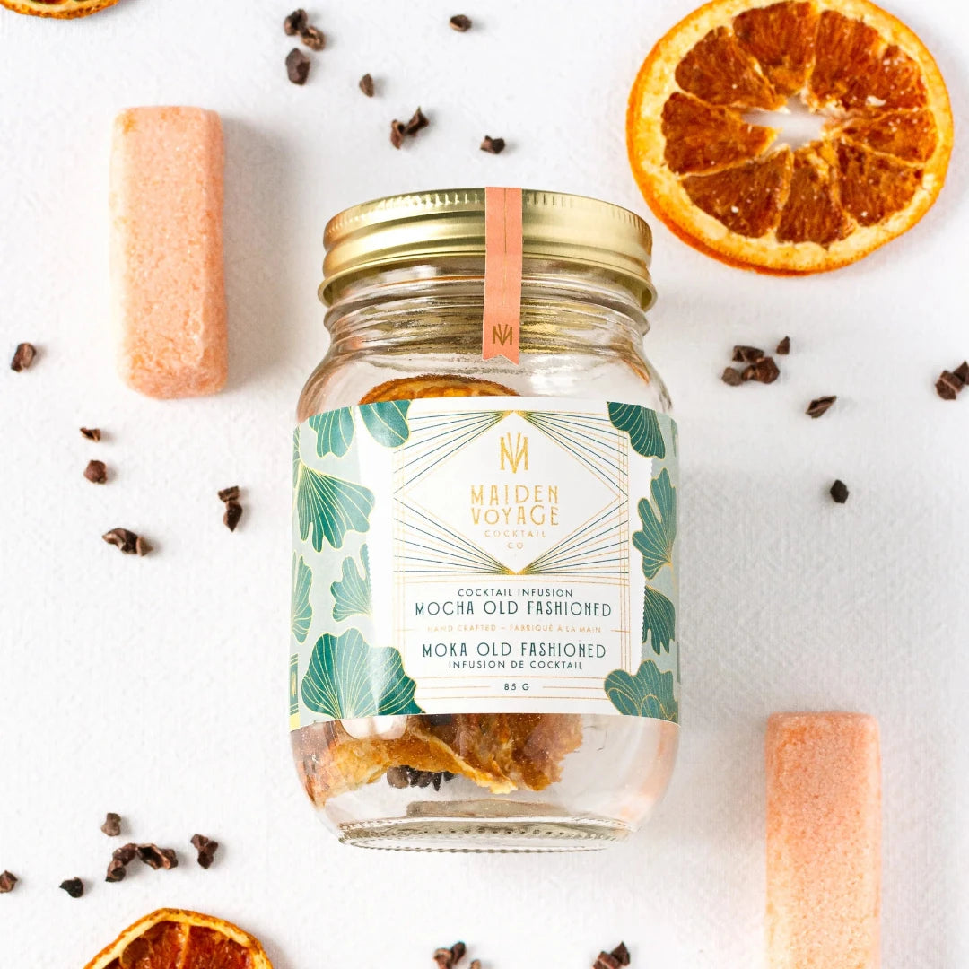 A glass jar filled with dried orange slices, cocoa nibs, and other ingredients. The jar has a white label with the Maiden Voyage logo and the product name "Mocha Old Fashioned Cocktail Infusion."
