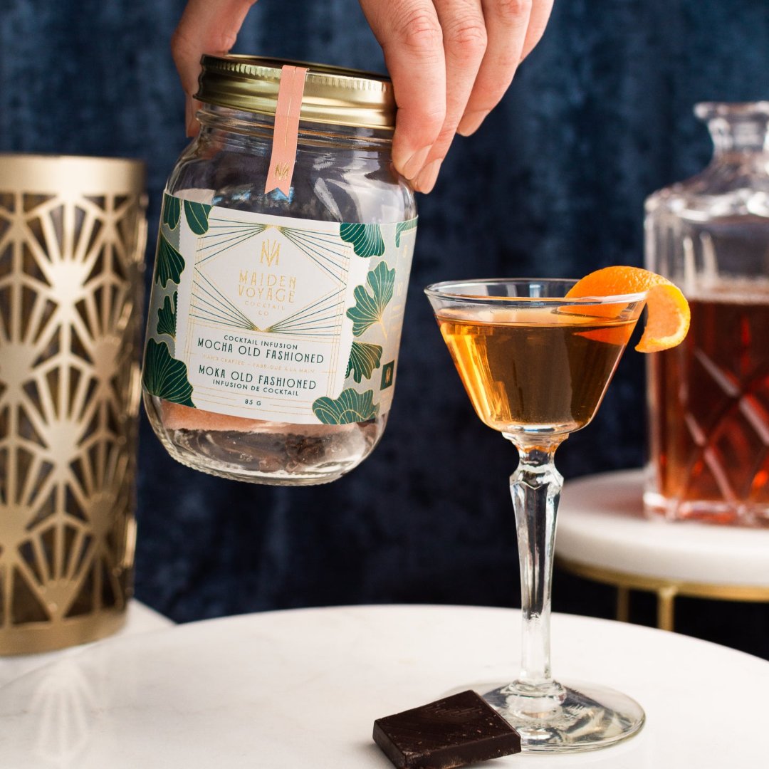 A glass jar filled with dried orange slices, cocoa nibs, and other ingredients. The jar has a white label with the Maiden Voyage logo and the product name "Mocha Old Fashioned Cocktail Infusion."