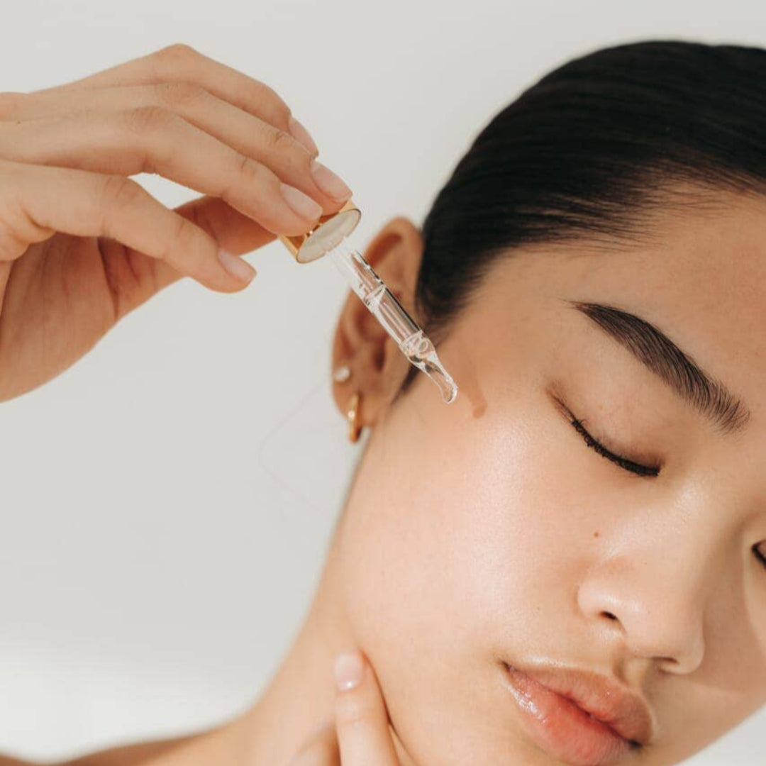 Person applying a dropper of skincare product to another person's face against a neutral background